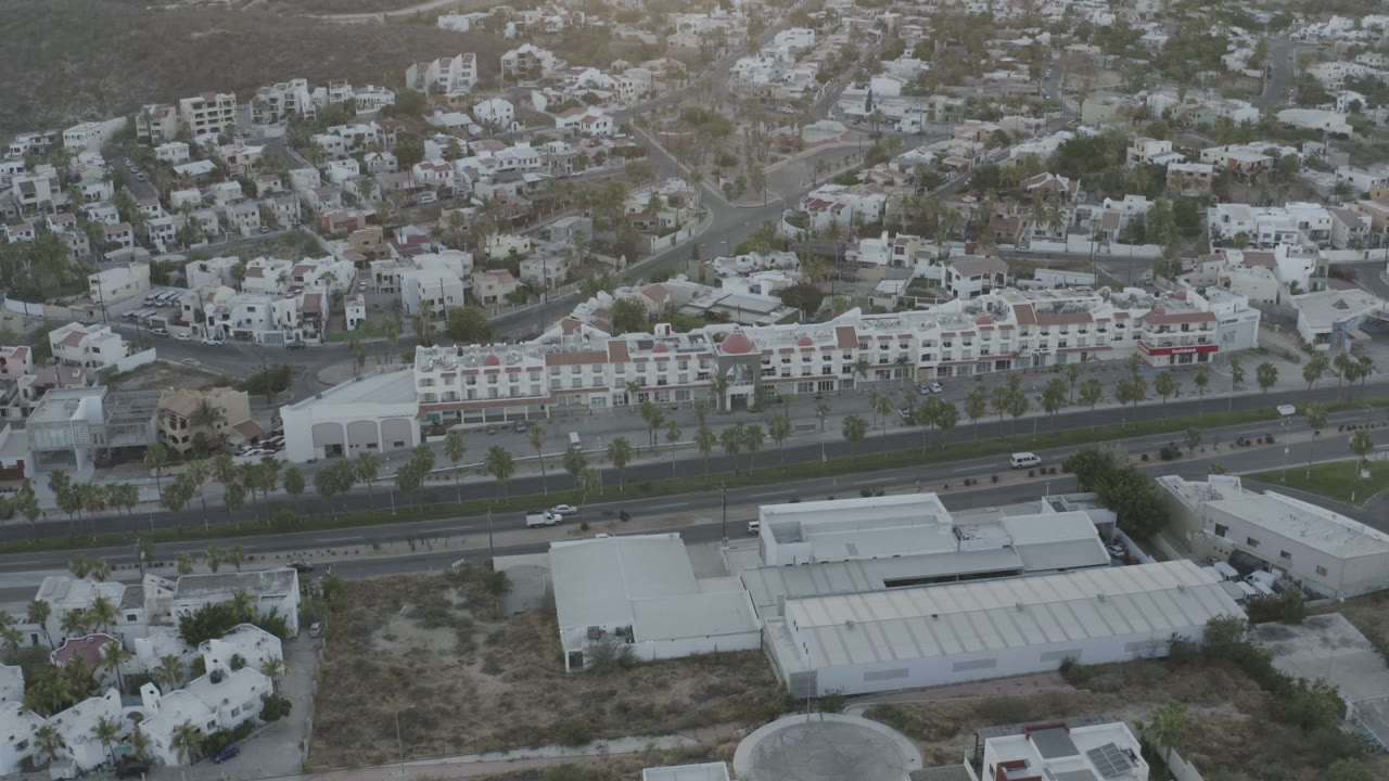 Second aerial frame over Los Cabos, Mexico, centering a long white building row, palm-lined road edges, hillside streets, and no visible people.