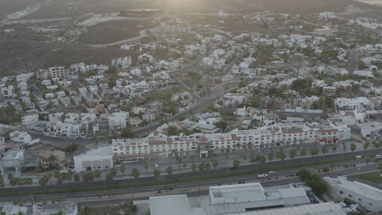 Aerial view over Los Cabos buildings and hillside roads with low sun glare near the top of frame and no visible people.