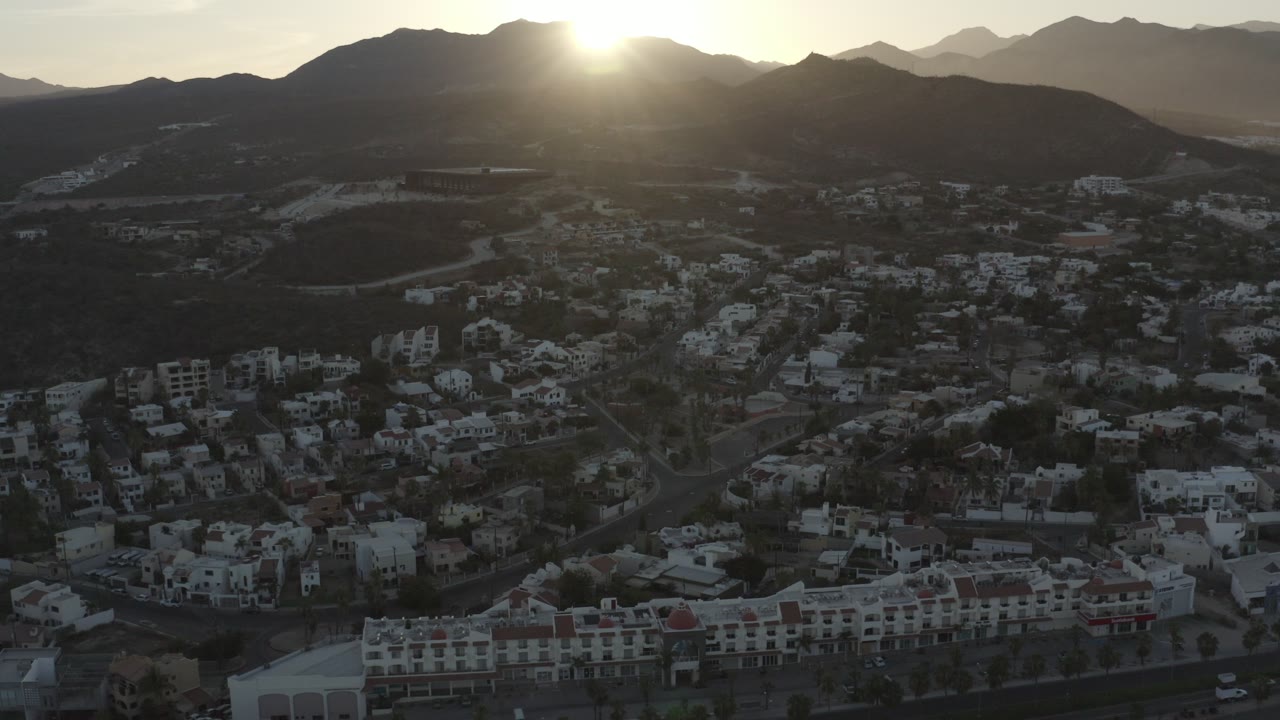 Wide aerial view over Los Cabos, Mexico, with hillside neighborhoods below a dark mountain ridge and the sun just above the horizon, no visible people.