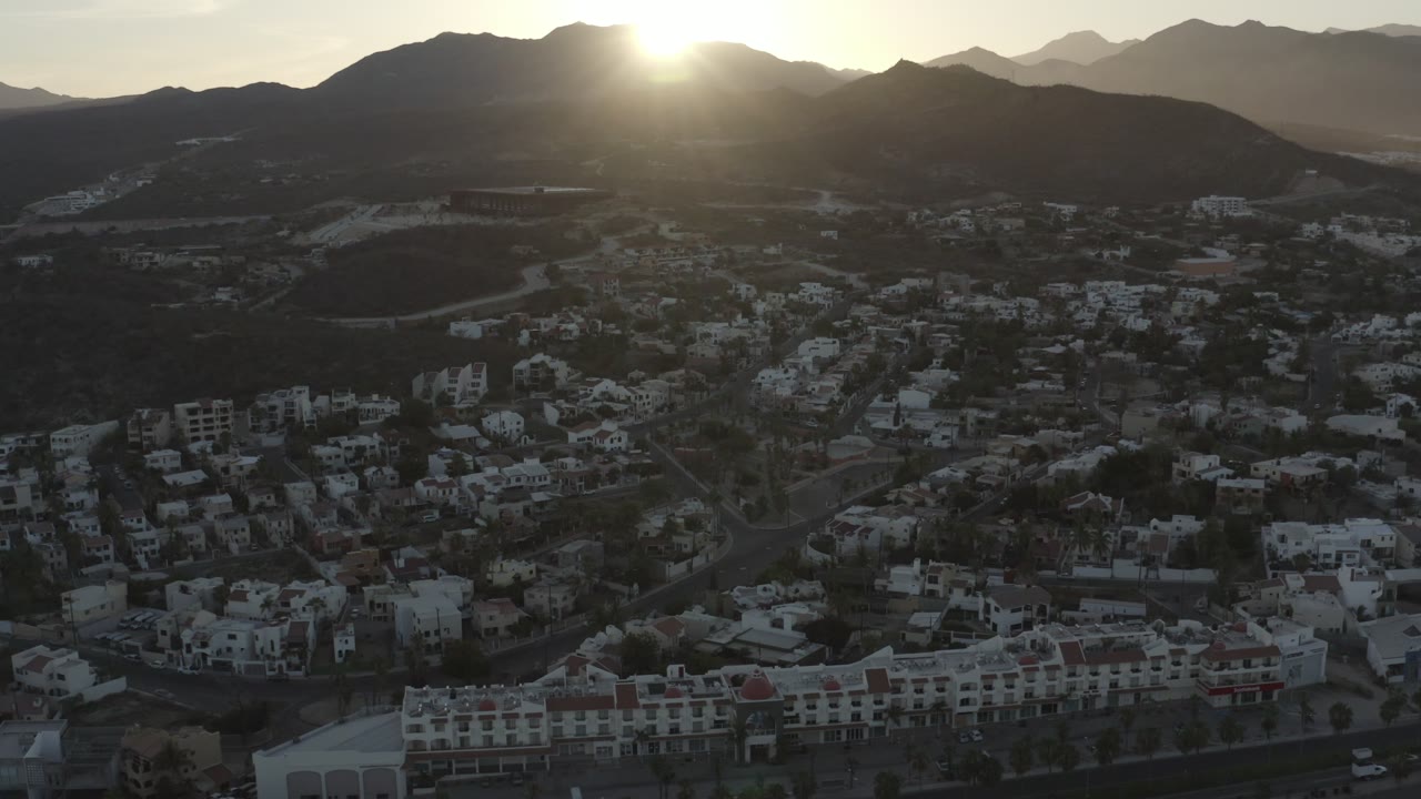 Los Cabos hillside buildings sit in deep shadow as the sun flares over the mountain ridge, with roads and residential blocks visible below, no visible people.