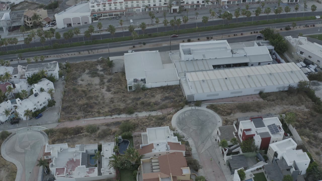 High overhead drone frame over Los Cabos showing a divided road at the top, white industrial roofs in the center, homes and curved streets below, and no visible people.