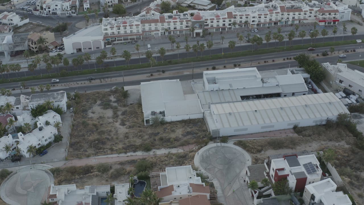 Opening aerial frame over Los Cabos, Mexico, with a divided road across the middle, pale rooftops below, low-rise buildings above, and no visible people.