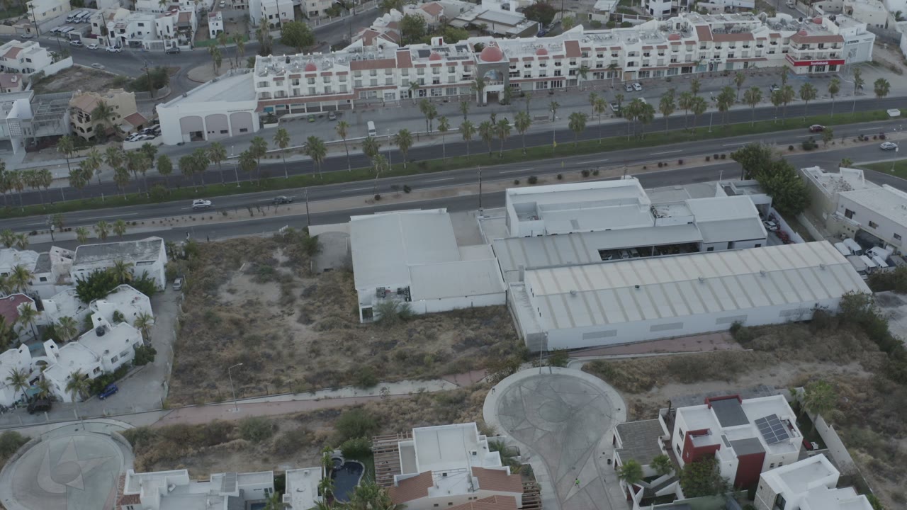 Drone-like view of a road, white buildings, palms, rooftops, and dry open ground.