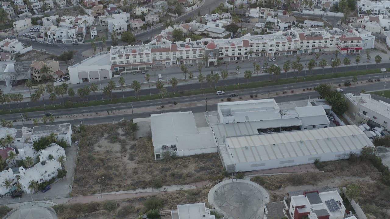 Los Cabos aerial view with a large pale roof near the lower center, a divided road with small cars, dense white housing beyond it, and no discernible people.