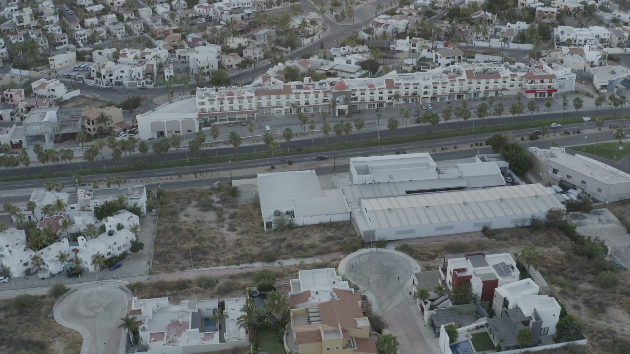 High aerial frame over Los Cabos, Mexico showing white residential buildings, dry open lots, a large roadway, hillside housing, and no visible people.