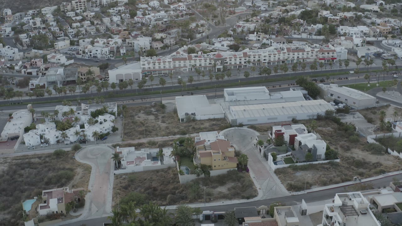 The Los Cabos aerial view centers on a white-roofed building, nearby villas, curving roads, dry parcels, and no visible people.