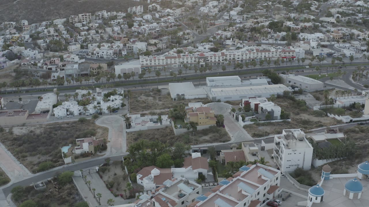Elevated Los Cabos view with white low-rise buildings at the bottom edge, a divided road through the middle, hillside housing above, and no visible people.