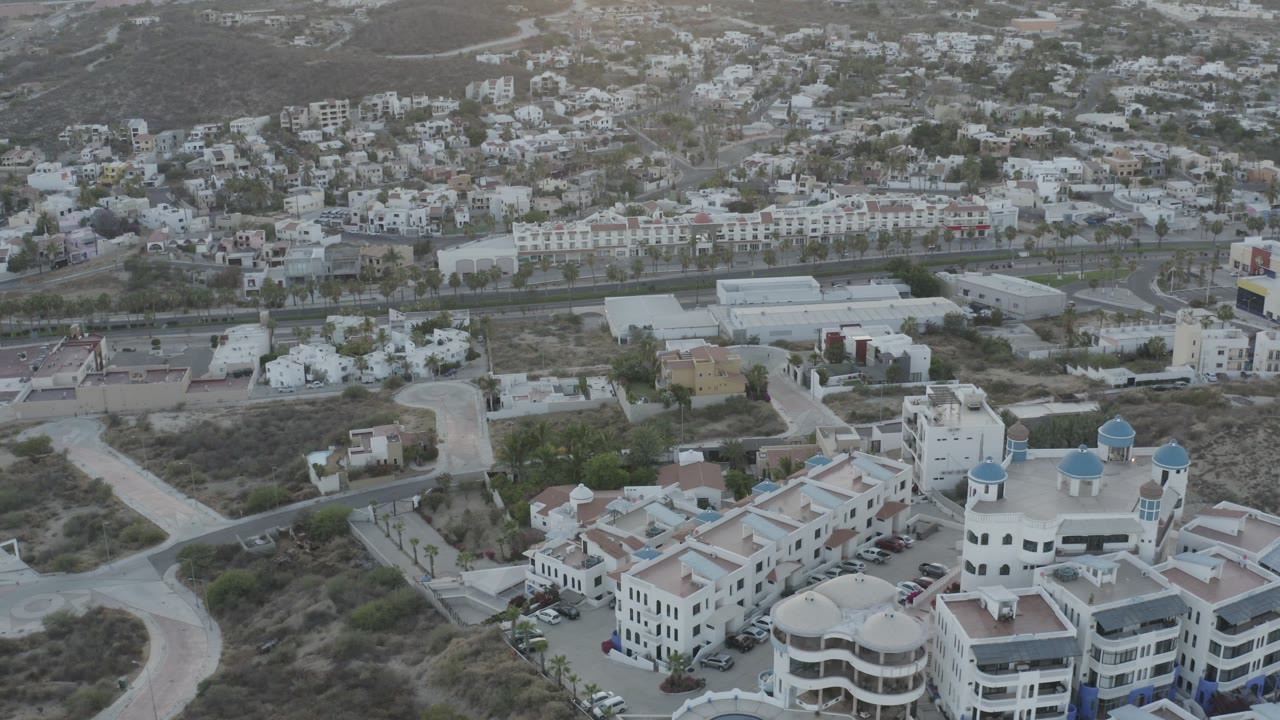 Aerial view in Los Cabos, Mexico, with the resort complex centered lower in frame, pale roads around dry lots, hillside homes beyond, and no visible people.