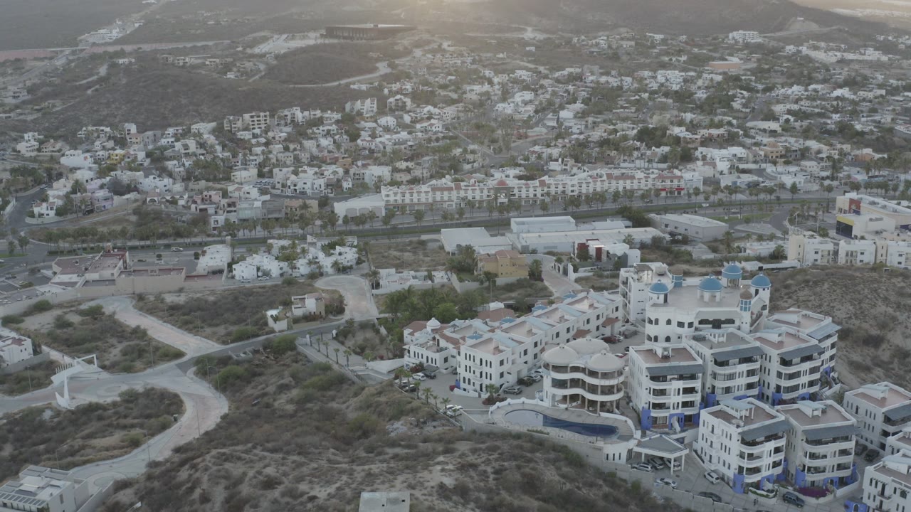 Aerial frame from Los Cabos showing clustered white homes and modern buildings on dry terrain, with a road network, distant water, mountains, and no visible people.