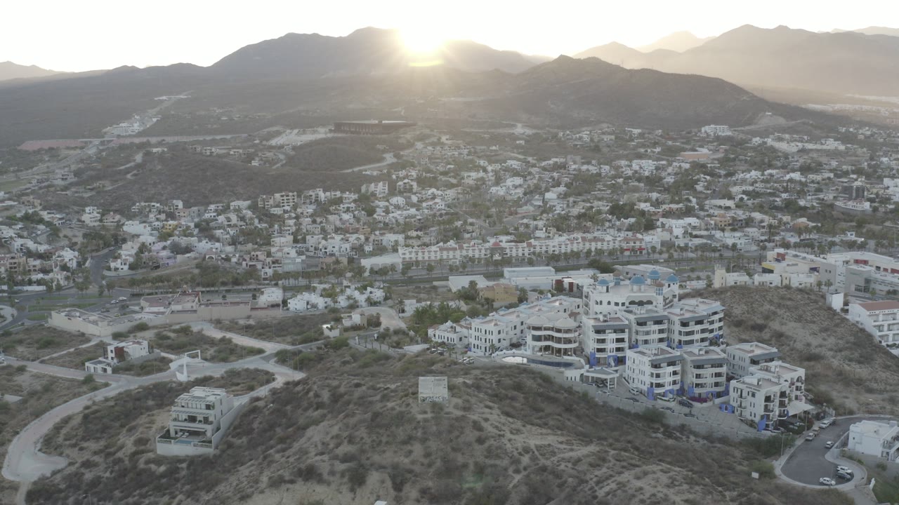 Aerial view in Los Cabos, Mexico showing white buildings on a dry hillside above valley roads, with mountains and intense sun glare in the background, no visible people.