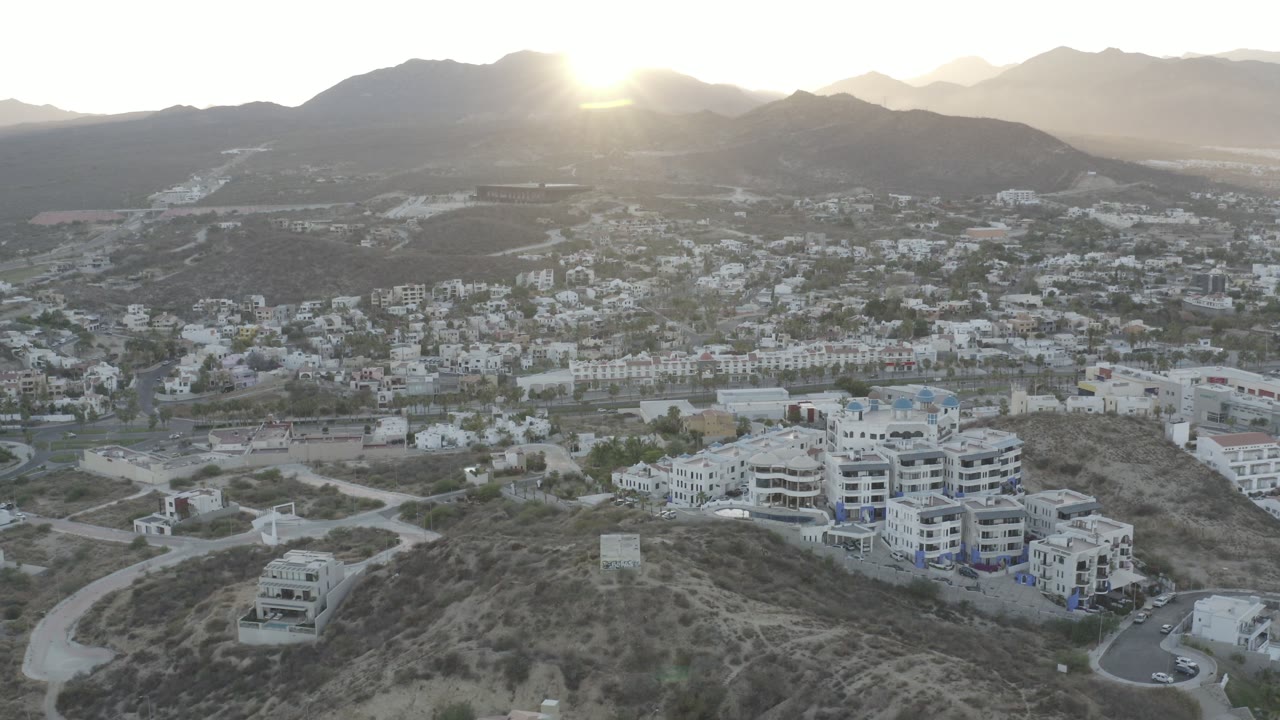Backlit aerial view over hillside roads, white buildings, a valley, and mountain ridges.