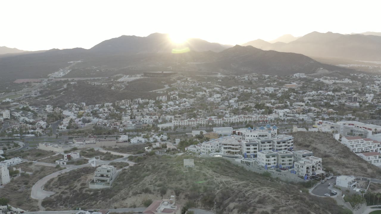 Opening frame of Los Cabos, Mexico, showing white hillside buildings above a valley town with mountains and low sun glare, with no visible people.