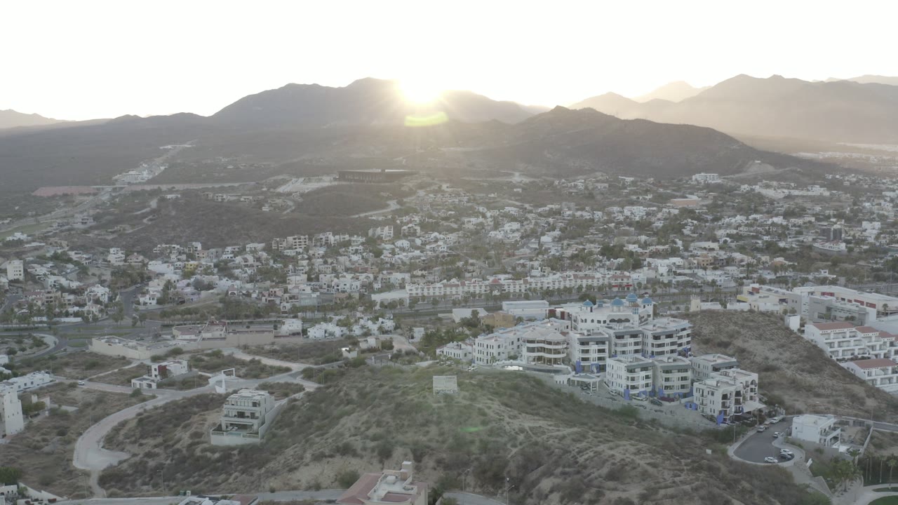 Los Cabos aerial frame with the sun sitting above dark mountain ridges, white residential blocks in the valley, and no visible people.