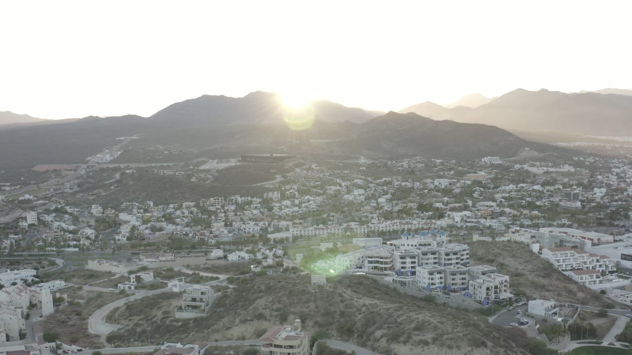 A high view over Los Cabos, Mexico, shows clustered light buildings across the hillside, a mountain ridge behind them, a bright sun near the ridge, and no visible people.