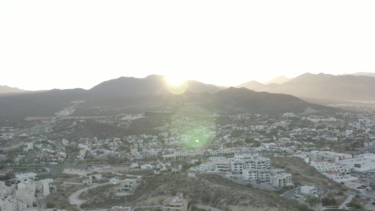 Wide aerial view over Los Cabos hillside homes and a desert valley, with the sun near the mountain ridge and no visible people.