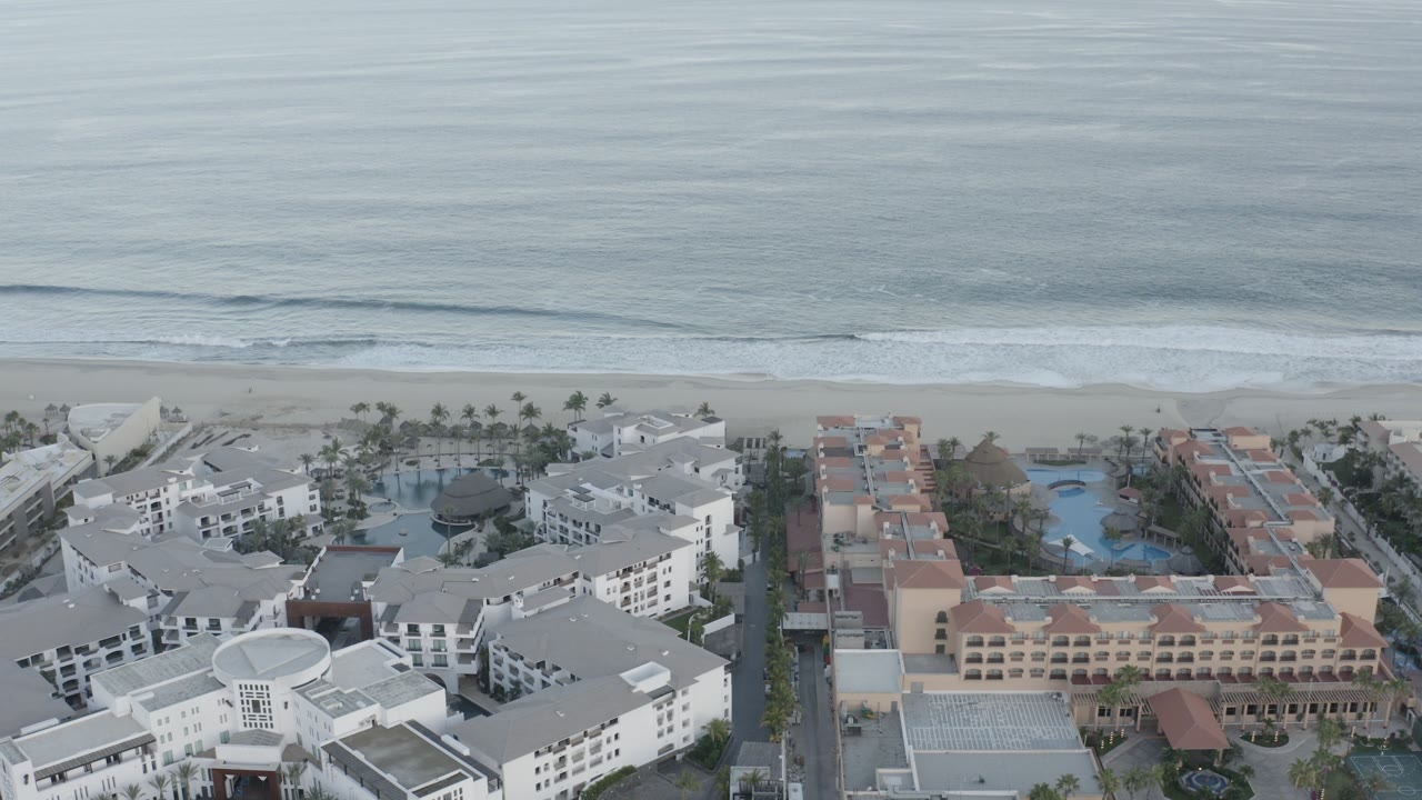 Aerial frame over Los Cabos resort-style buildings and pools, with the shoreline running across the middle and no recognizable people visible.