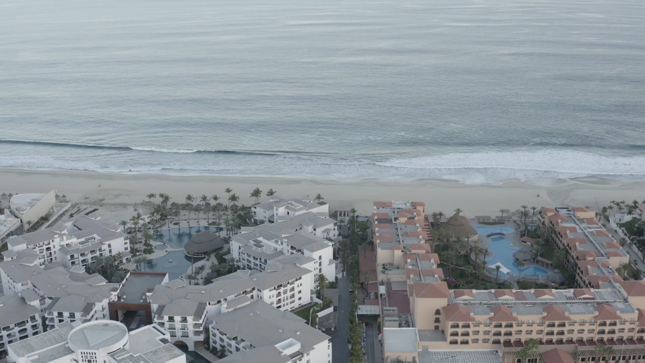 Aerial frame in Los Cabos, Mexico, showing rolling surf, a pale beach, palm-lined pools, white apartment blocks, and peach-roofed buildings, with no visible people.