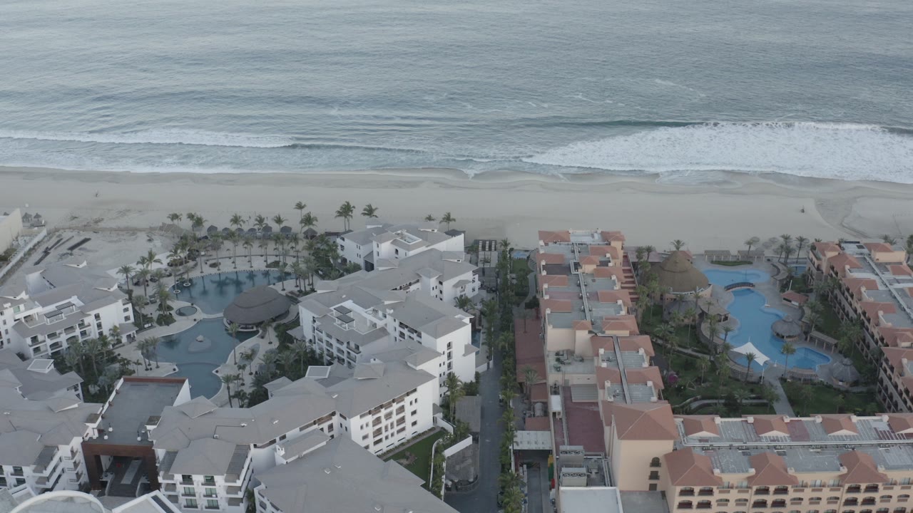 Opening aerial frame over Los Cabos resort buildings with pools and palm landscaping below a pale beach and gray-blue surf, with no visible people.