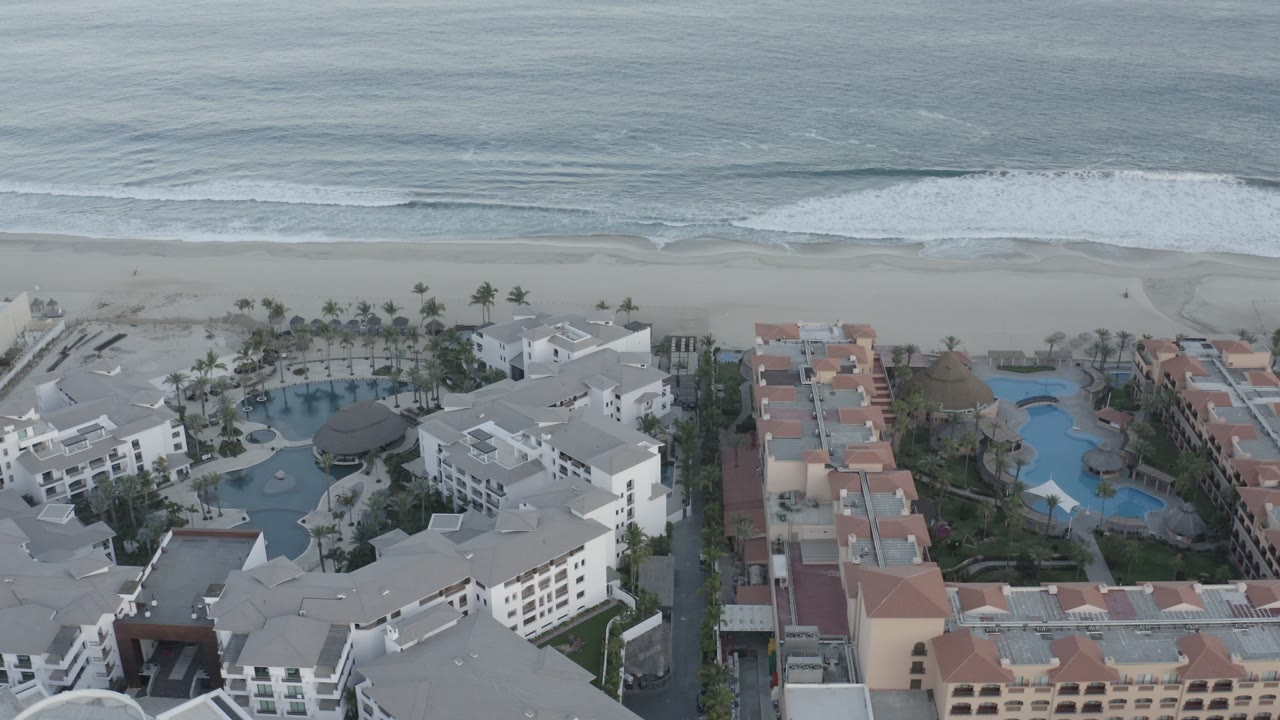 Los Cabos aerial view with white resort buildings in the foreground, a curved pool area to the left, sandy beach beyond, and no visible people.