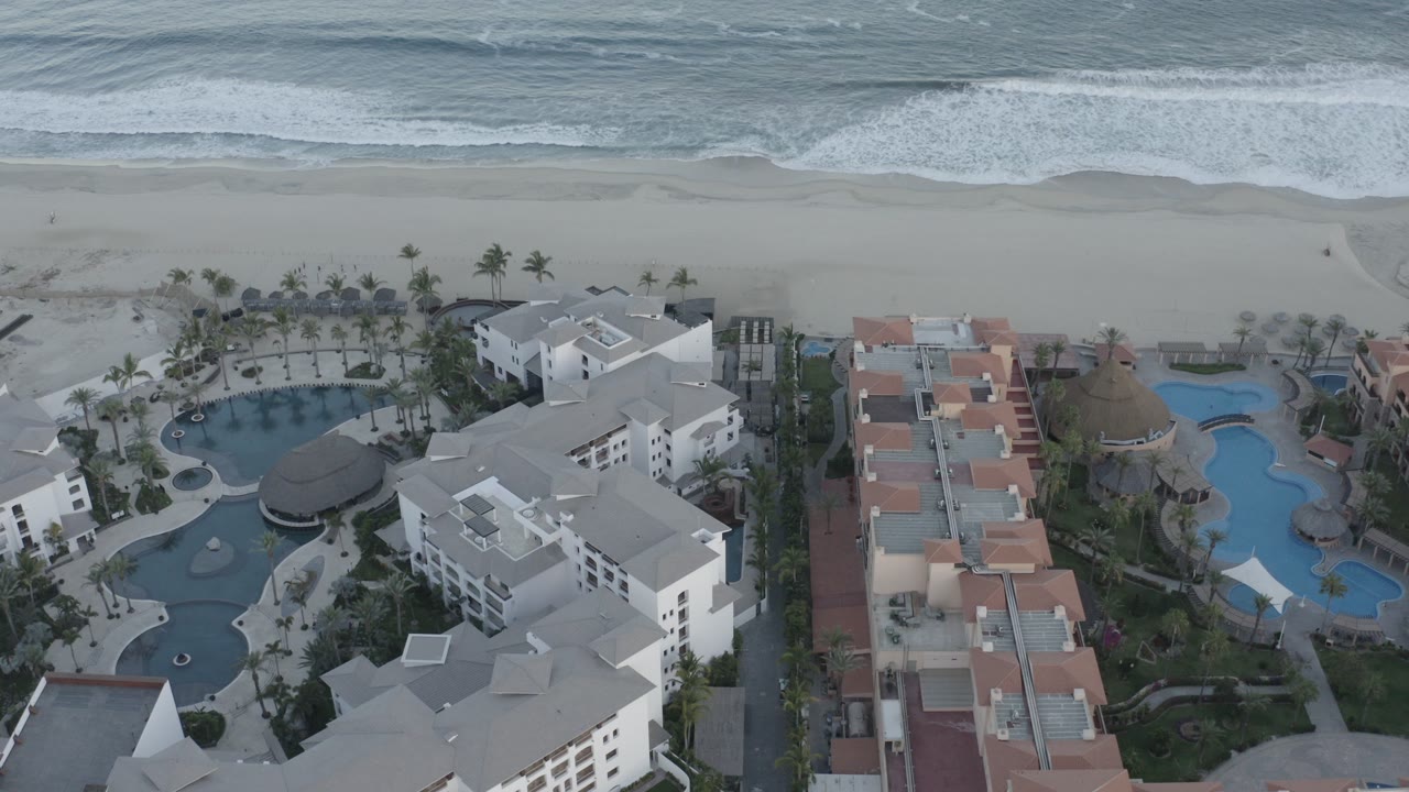 Opening aerial frame over Los Cabos resort buildings with curving blue pools below, pale beach and gray-blue surf beyond, and no visible people.