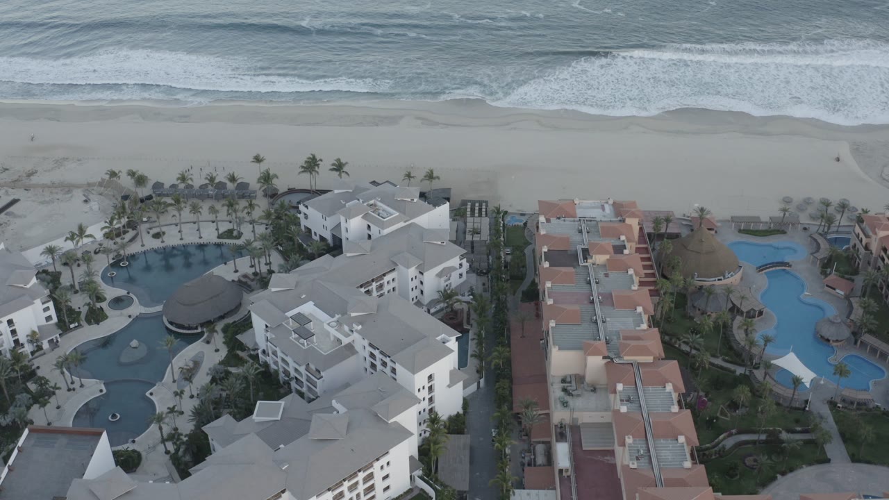 Aerial frame in Los Cabos showing white multi-story buildings flanked by large swimming pools, with a wide beach and breaking waves across the top, and no visible people.