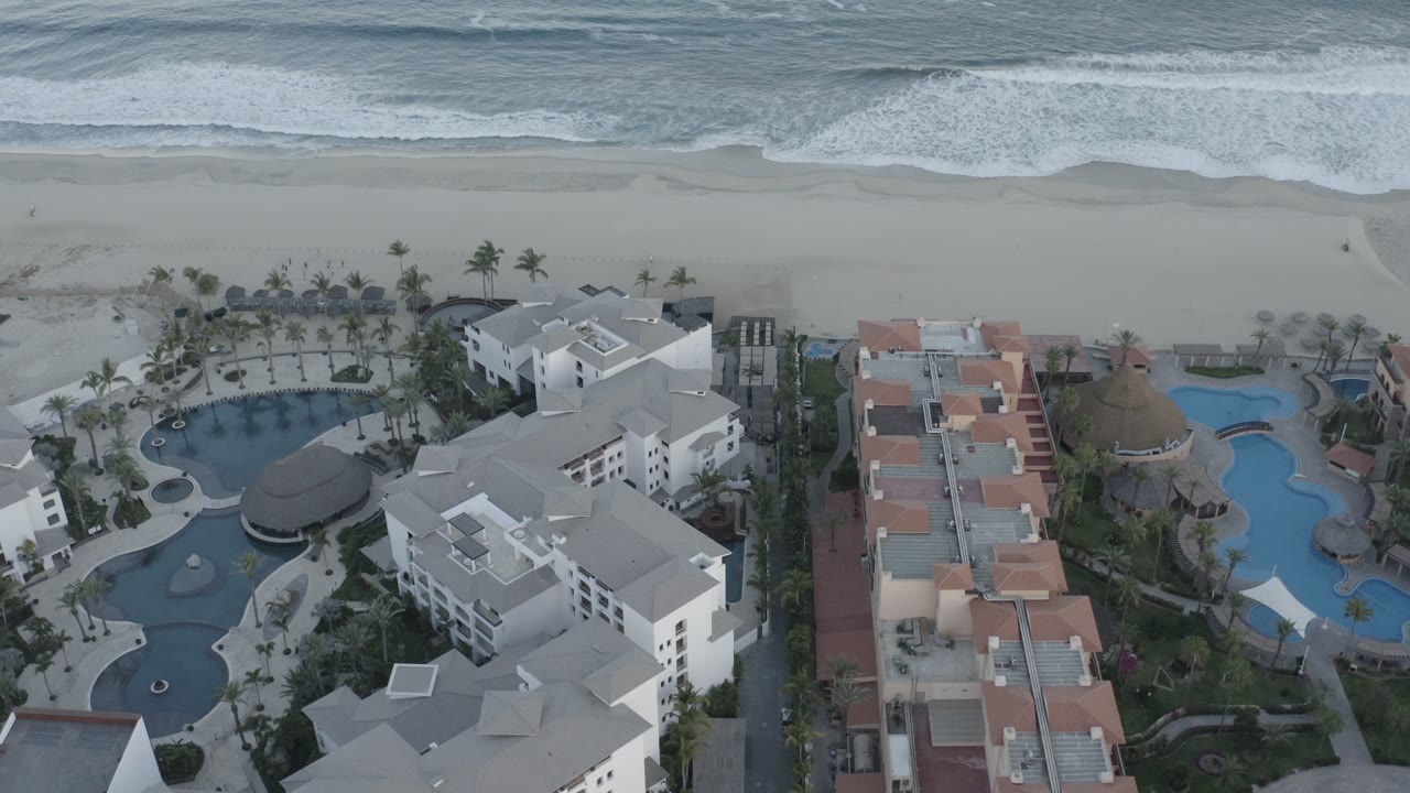 Drone-style view of resort roofs, pools, palm trees, beach, and sea.