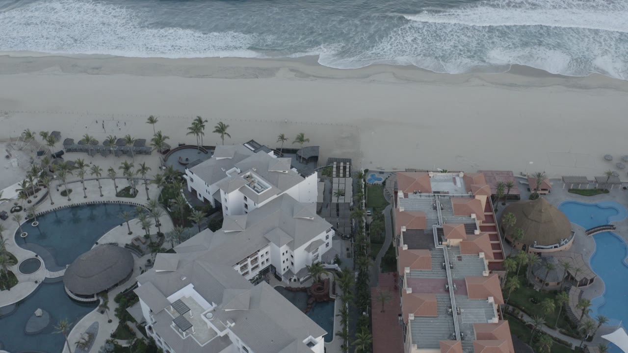 High aerial frame over a Los Cabos resort with white buildings, curved pools, palm rows, pale beach, breaking waves, and no visible people.