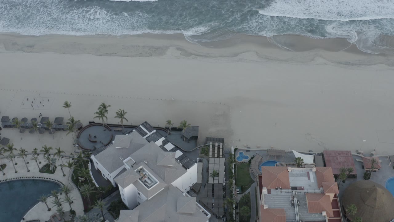 Overhead beach view with surf, wet sand, hotel roofs, pools, and palm trees.