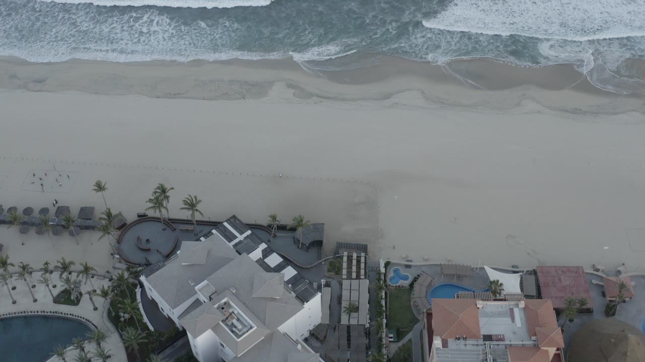 Top-down shoreline with foamy water, smooth sand, palm-lined hotel grounds, and pools.