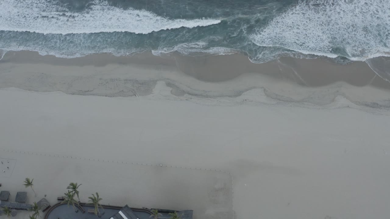 Aerial frame of white surf breaking across the upper edge of a pale Los Cabos beach, with faint sand texture and no visible people.