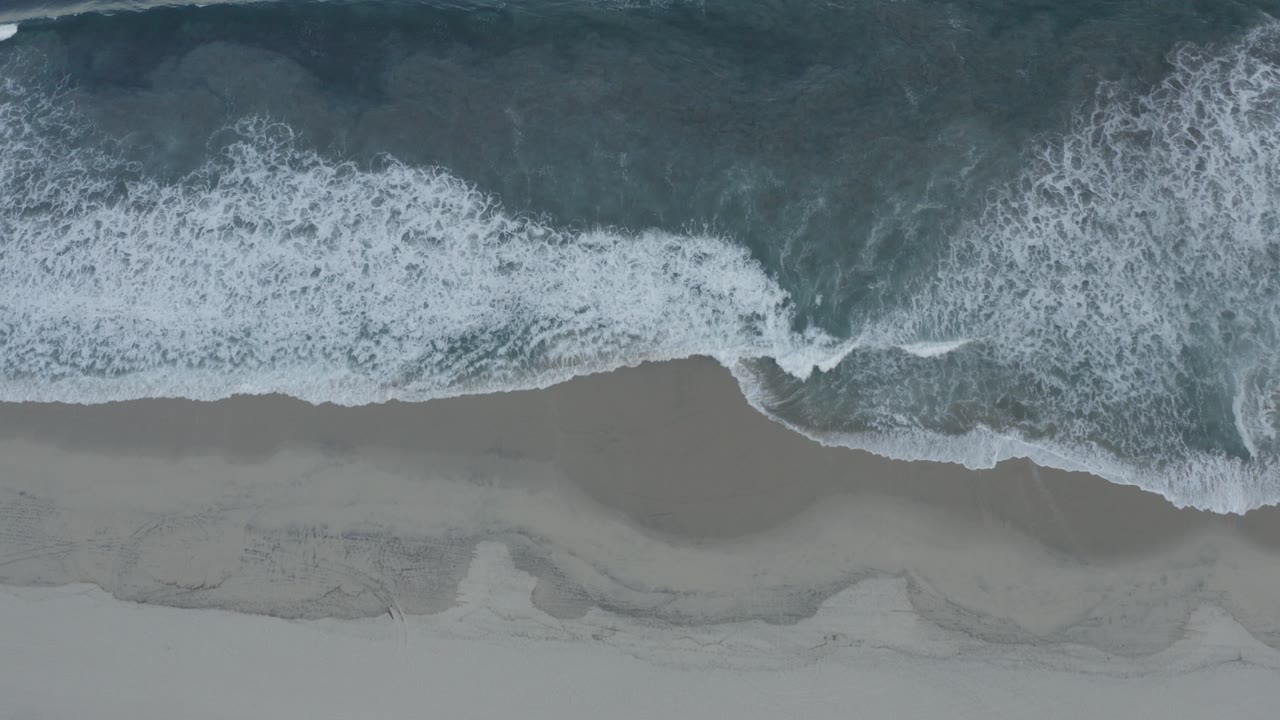 Top-down aerial view of Los Cabos ocean surf spreading white foam onto gray-tan sand, with no visible people.