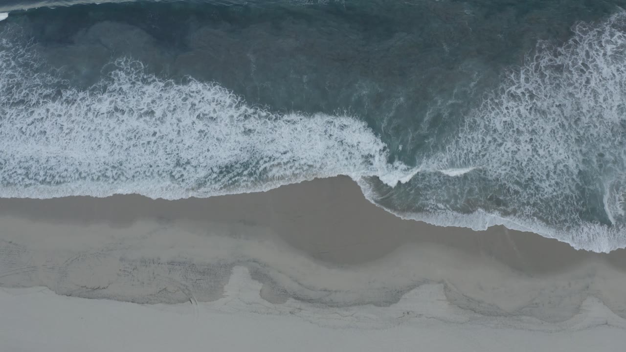 A wider band of white surf crosses the Los Cabos shoreline from above, with darker water above and empty sand below.