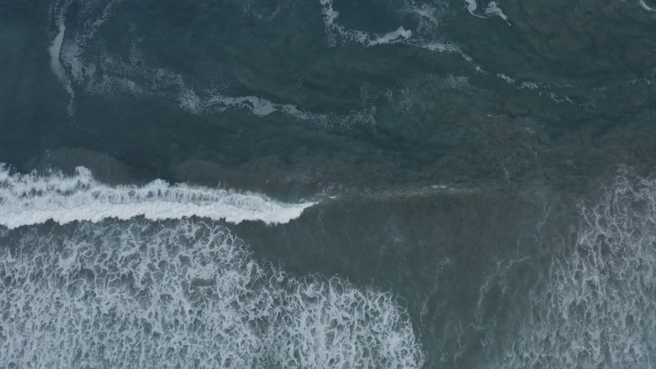 Top-down aerial view of dark blue-green ocean in Los Cabos, Mexico, with a bright white wave breaking across the lower part of the frame and no visible people.