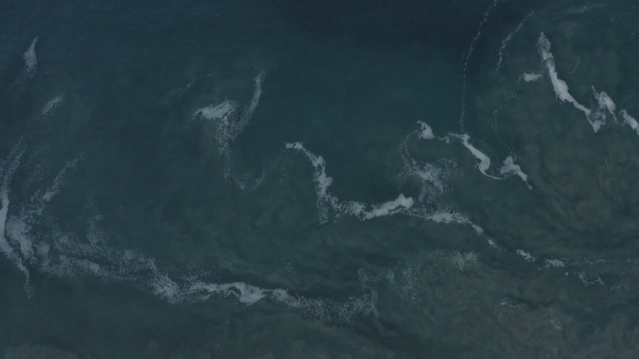 Top-down aerial view of dark ocean water in Los Cabos, Mexico, with scattered pale foam streaks near the upper and lower edges and no visible people.