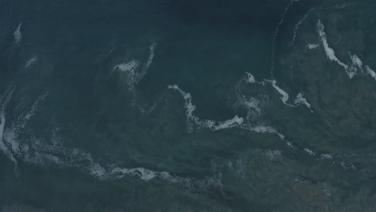 Aerial frame over dark blue-green water in Los Cabos, Mexico, with curved white foam marks crossing the upper half and no visible people.