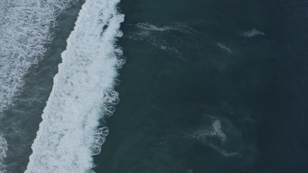 Top-down view of blue-green ocean in Los Cabos, Mexico, with a thick diagonal line of breaking white surf and no visible people.