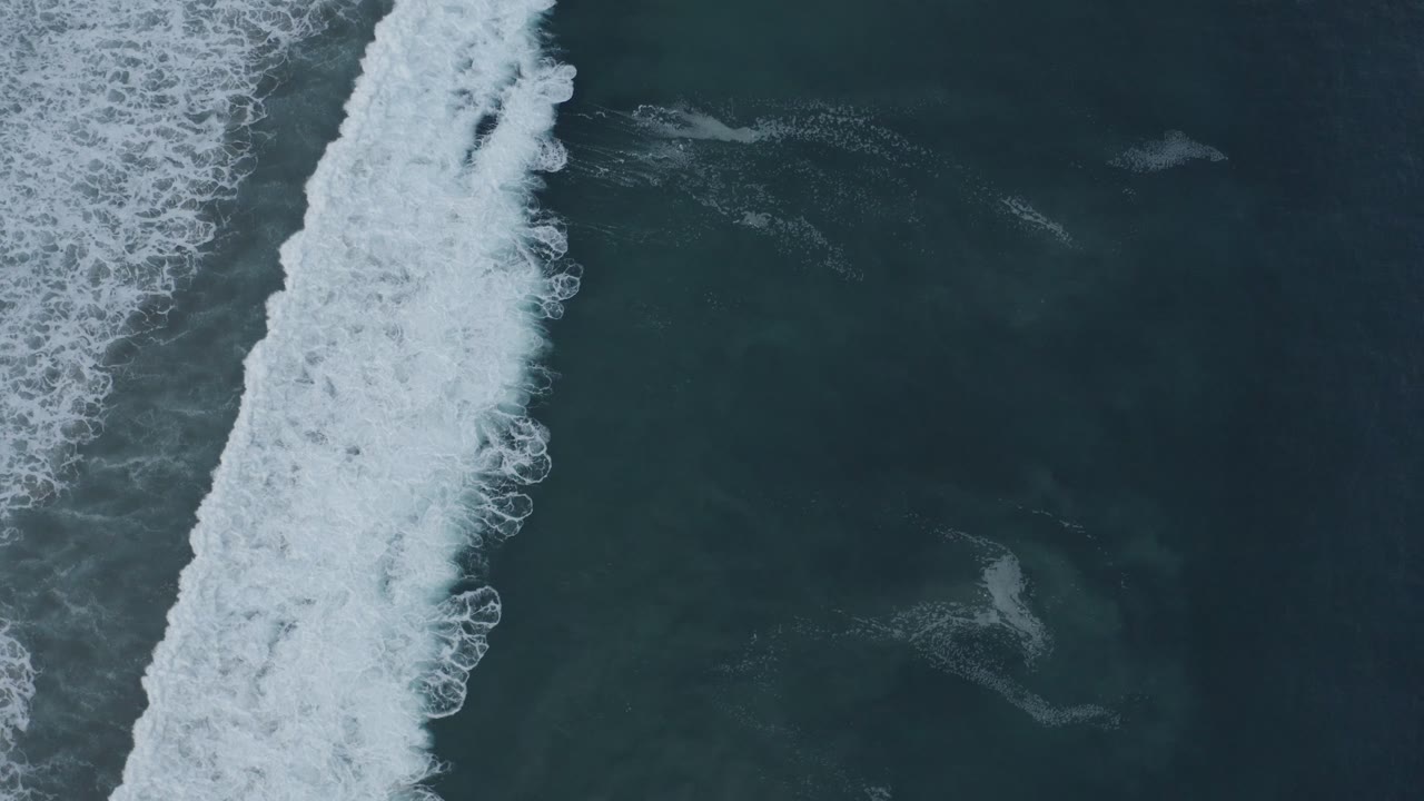 Aerial frame in Los Cabos, Mexico, showing white foam advancing across darker blue water with no visible people or boats.