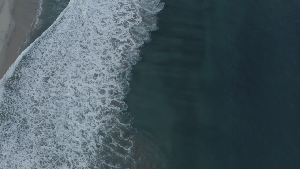 Aerial Los Cabos shoreline frame with dense white foam widening across the center and a muted strip of beach along the left edge, no visible people.