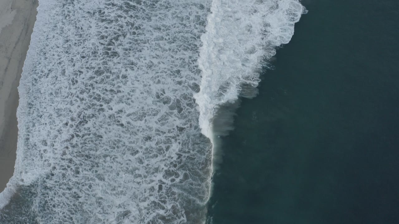 Overhead view in Los Cabos, Mexico, with a wide white surf band breaking beside wet gray sand and no visible people.