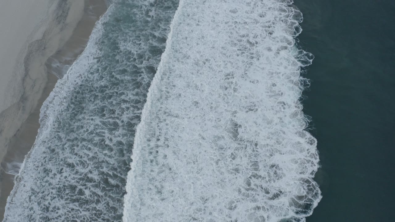 Overhead aerial view in Los Cabos, Mexico, showing white surf breaking along dark teal water and gray-tan wet sand with no visible people.