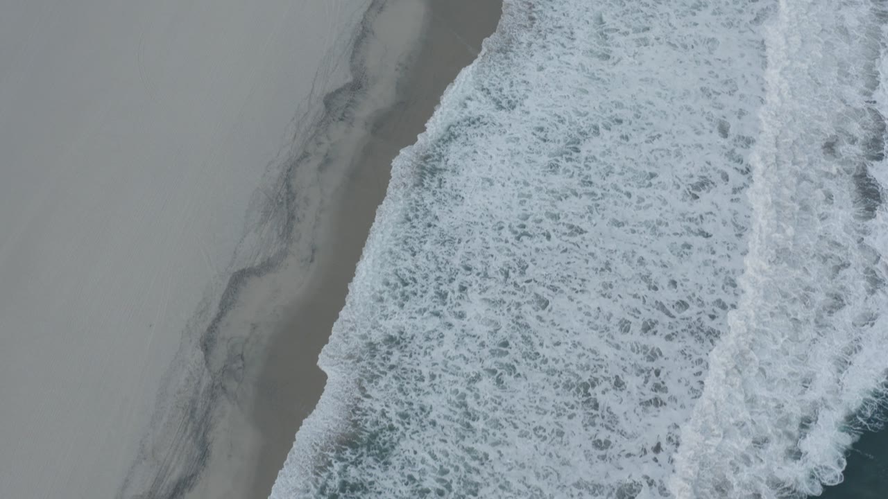 Overhead view of white surf meeting pale sand along a diagonal shoreline.