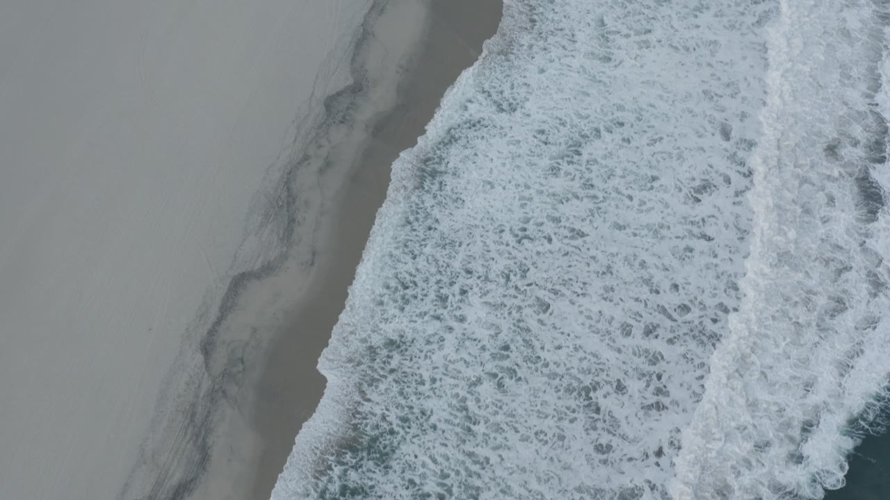 Aerial view in Los Cabos showing dense white foam advancing beside a dark gray wet-sand edge, with broad empty sand to the left and no visible people.