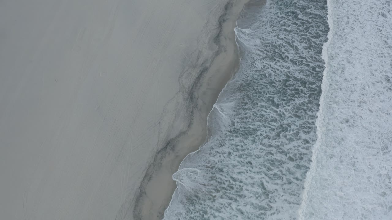 Aerial Los Cabos shoreline frame with a broad strip of pale beach, dark water marks near the surf edge, white foam, and no visible people.