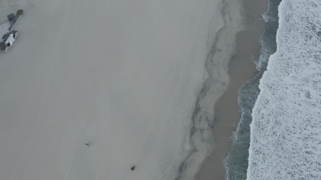 Aerial view of the Los Cabos shoreline with surf pressing in from the right, pale open sand through the middle, faint tracks, and no visible people.