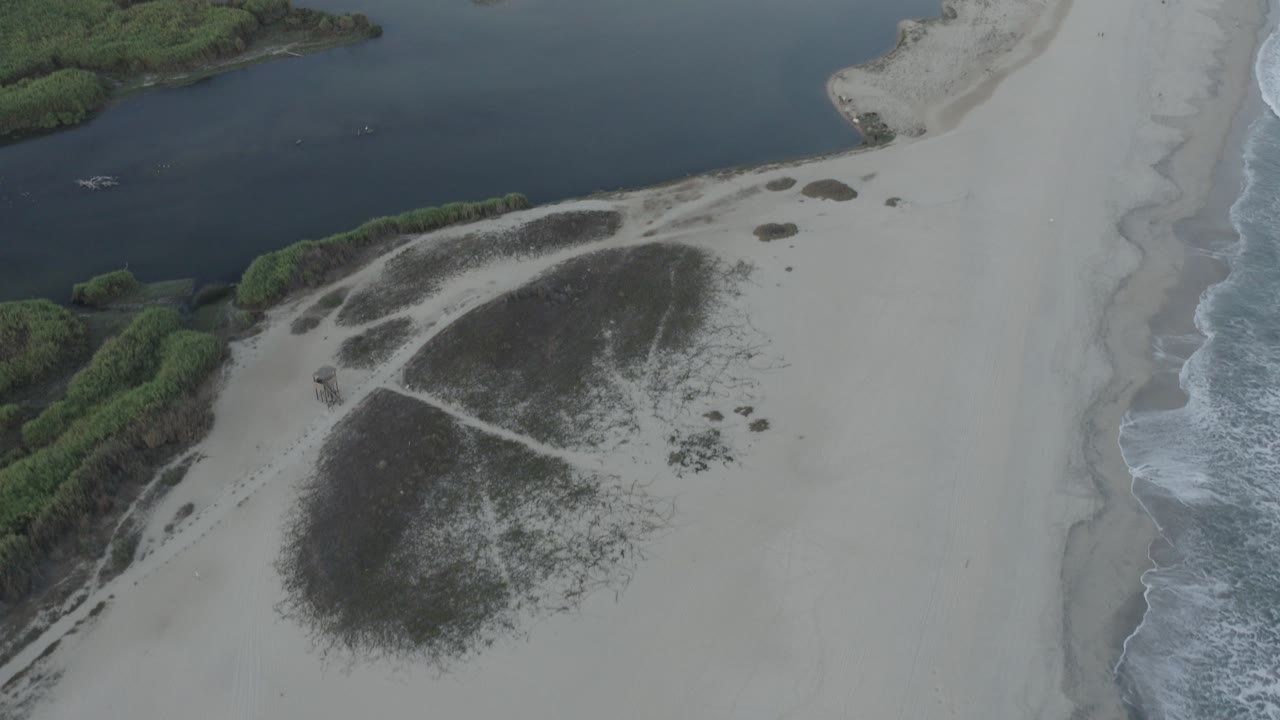 Aerial view in Los Cabos, Mexico, with dark lagoon water above pale sand, low green shrubs at the edge, surf on the right, and no visible people.