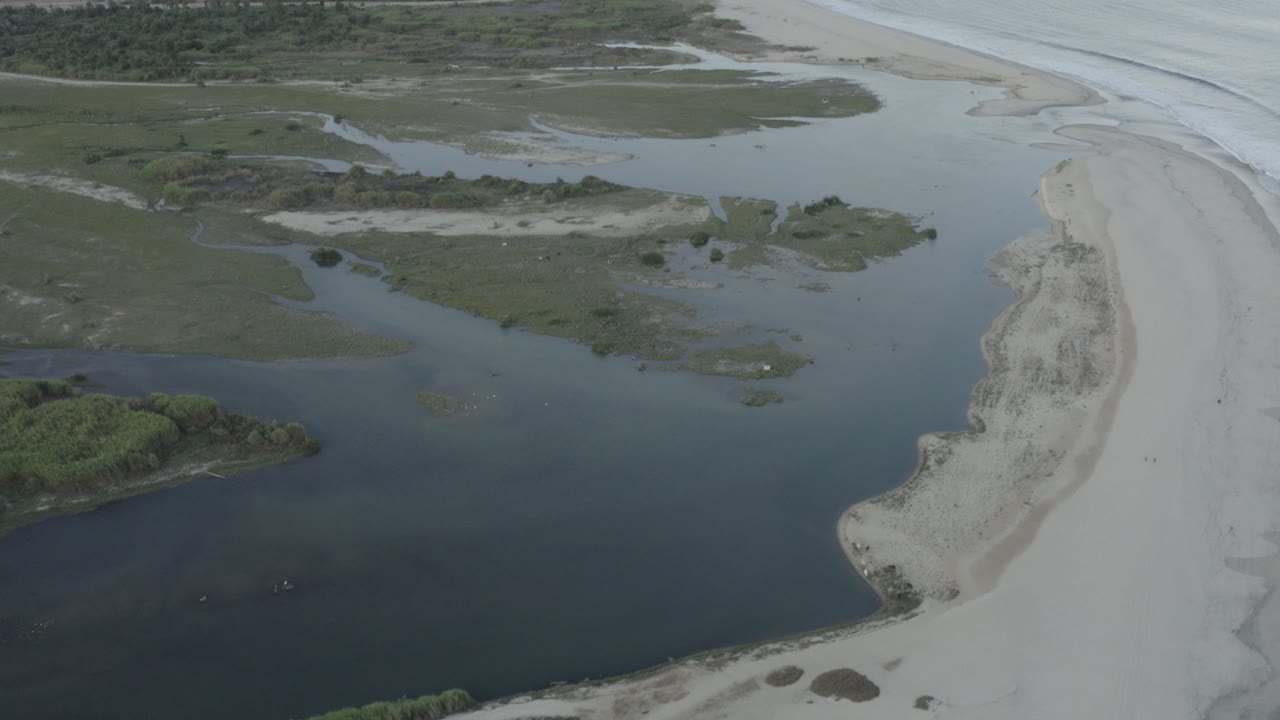 High aerial view over Los Cabos, Mexico, with dark inlet water in the foreground, a pale sand beach at right, ocean surf beyond it, green vegetation inland, and no visible people.