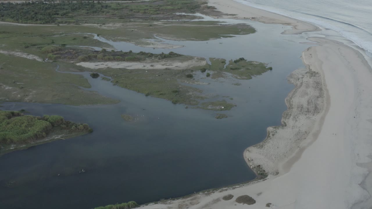 A Los Cabos aerial frame shows lagoon-like inlet channels across green coastal vegetation, a narrow sandy shoreline to the right, ocean waves near the top edge, and no visible people.