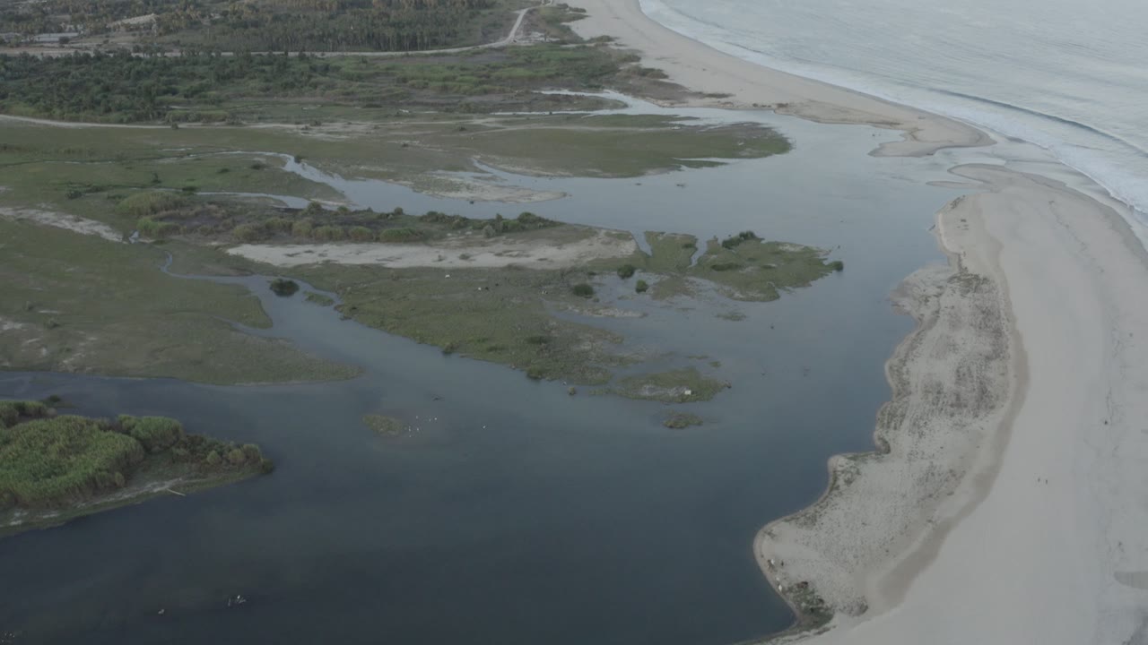Wide aerial coast with inlet water, sand, surf, vegetation, and distant roads.
