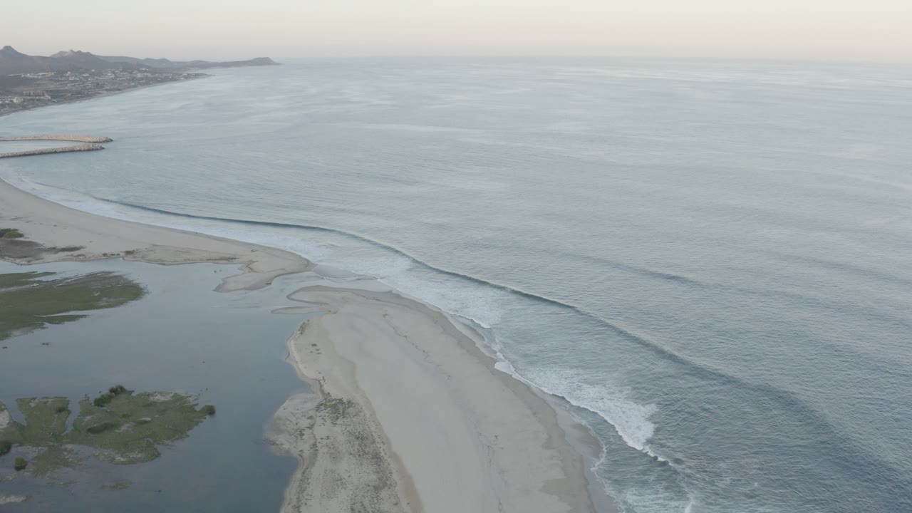 Aerial Los Cabos coastline frame with pale sand in the lower center, soft surf bands along the beach, and distant hills under hazy light, with no visible people.