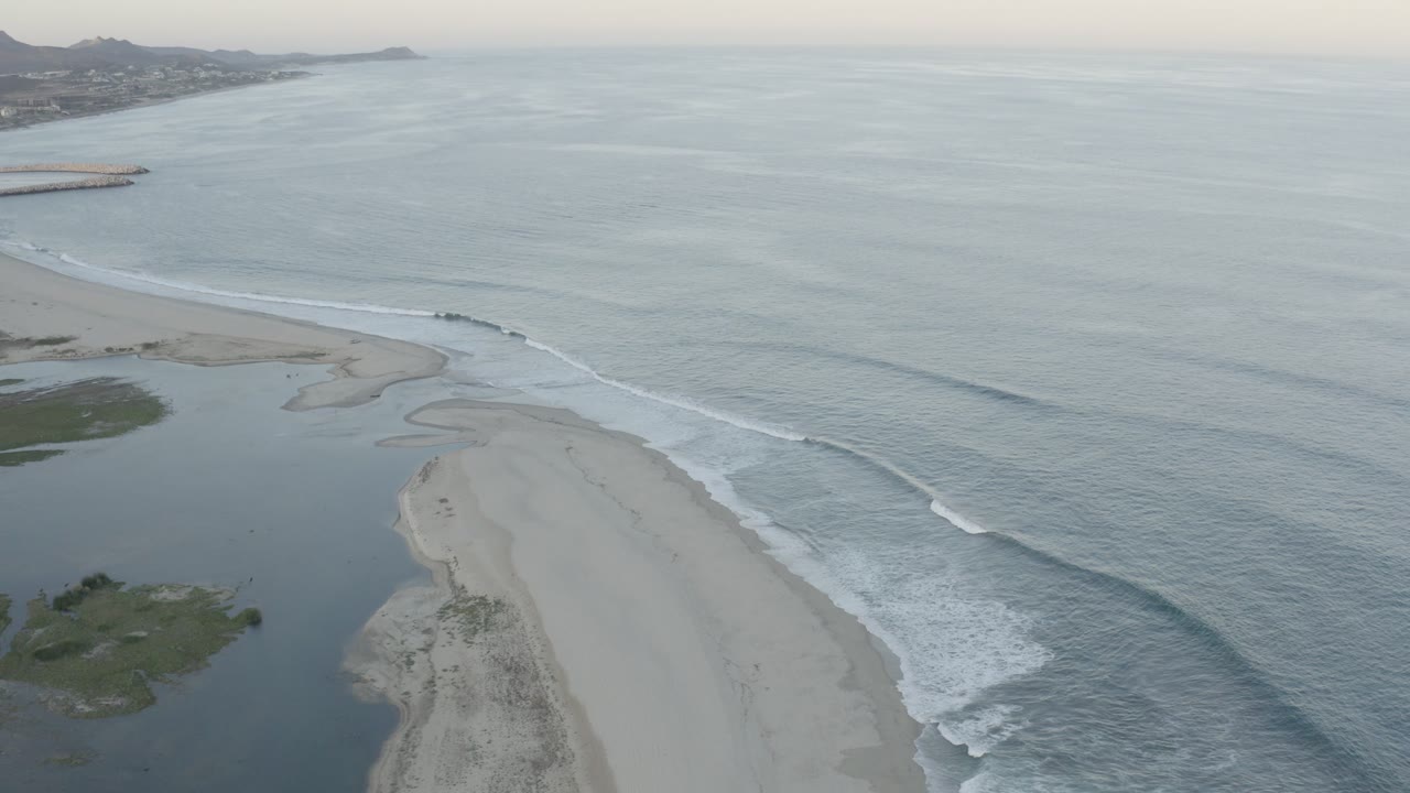 Aerial view of surf breaking along a sandy beach beside a dark lagoon.